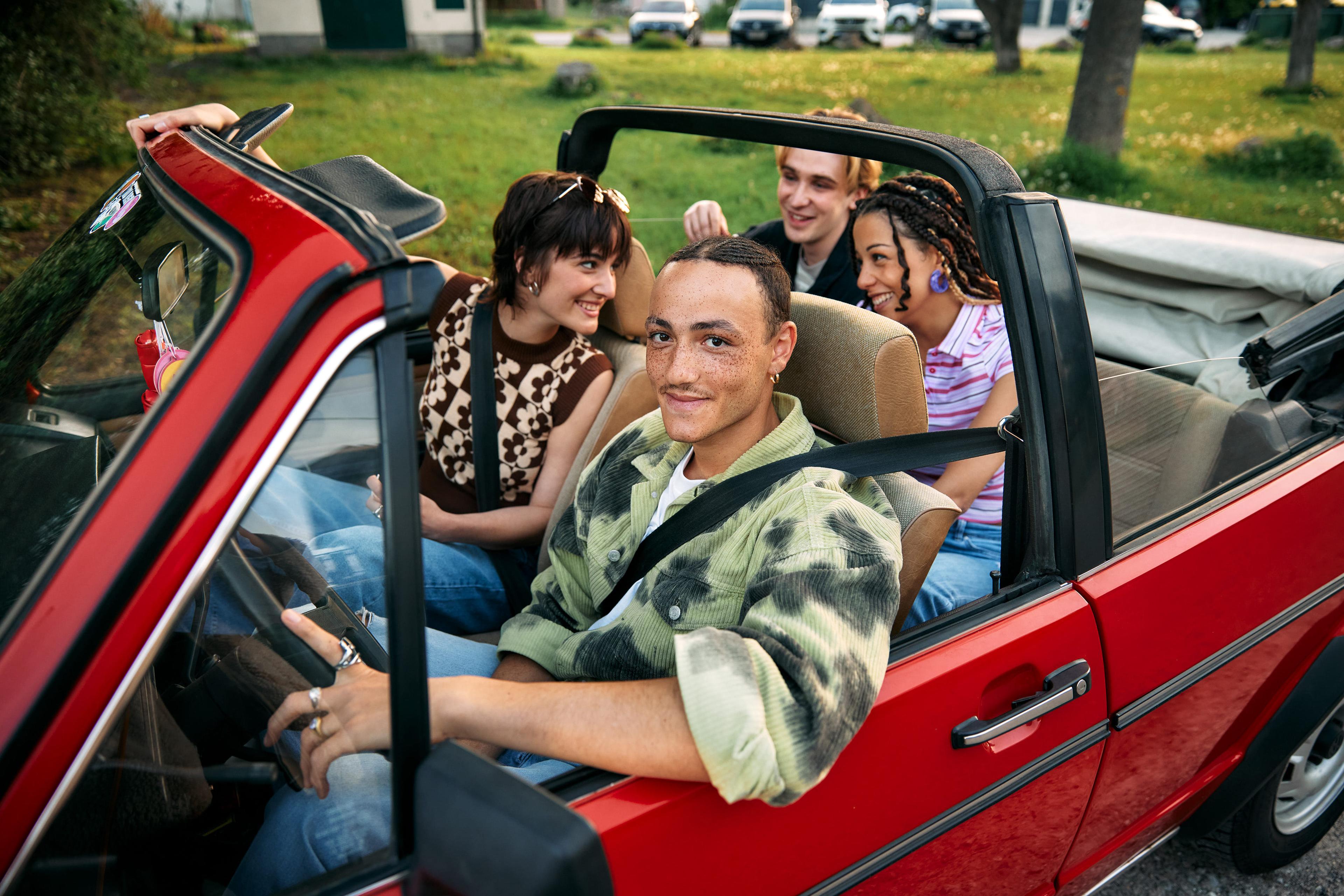 Zu sehen ist ein rotes Cabrio und vier Jugendliche. Während der Fahrer schon angeschnallt auf seinem Platz sitz, sind die anderen dabei, voller Vorfreude einzusteigen.