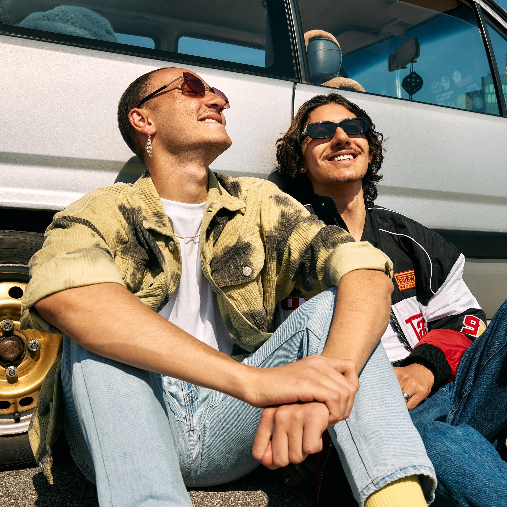 Zwei männliche Jugendliche machen eine Pause von der Autofahrt und sitzen entspannt auf dem Boden, mit dem Rücken an ihr Auto gelehnt. Sie schauen in die Sonne und lächeln freundlich.